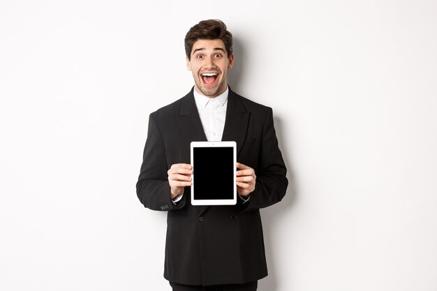 Image of attractive male entrepreneur in trendy suit, showing digital tablet screen and smiling amazed, standing over white background.