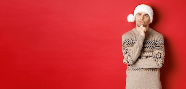 Image of attractive man in christmas sweater and santa hat, thinking about new year gifts, looking at upper left corner thoughtful, standing over red background.