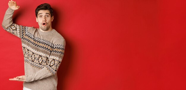 Image of attractive man in christmas sweater shaping big new year gift showing something large and amazing standing over red background