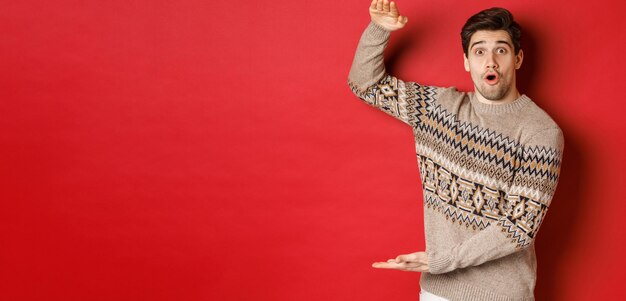 Image of attractive man in christmas sweater, shaping big new year gift, showing something large and amazing, standing over red background.