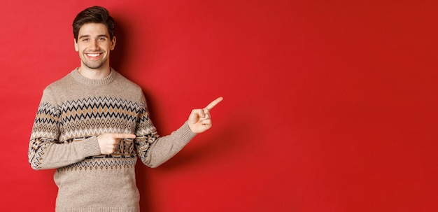 Image of attractive smiling man, wearing christmas sweater, pointing fingers right and showing new year advertisement, standing over red background.