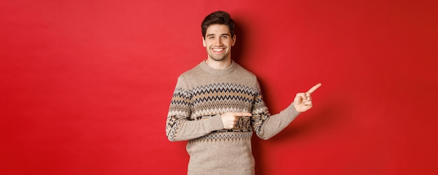 Image of attractive smiling man, wearing christmas sweater, pointing fingers right and showing new year advertisement, standing over red background.