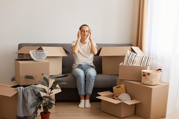 Image of attractive tired woman wearing white t shirt and jeans sitting on sofa during relocating to a new apartment being exhausted and trying to concentrate