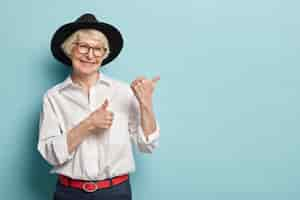 Free photo image of attractive wrinkled woman with appealing look, feels refreshed, young for her age, points at upper right corner, satisfied with product, wears white shirt, black headgear, optical glasses