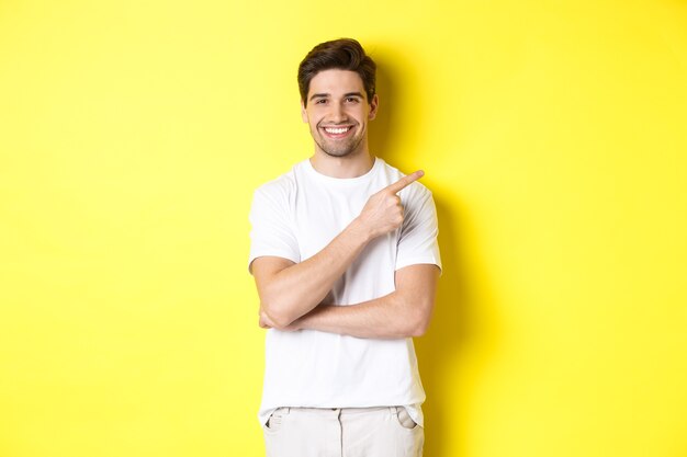 노란색 배경 위에 서 배너 또는 프로 모션 제안을 보여주는 복사 공간에서 오른쪽 손가락을 가리키는 매력적인 젊은 남자의 이미지.