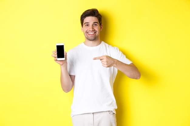 Image of attractive young man pointing finger at smartphone screen, showing an app, standing against yellow background