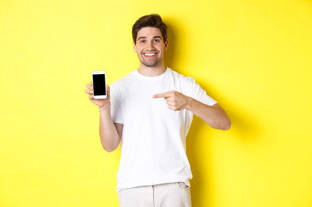 Image of attractive young man pointing finger at smartphone screen, showing an app, standing against yellow background