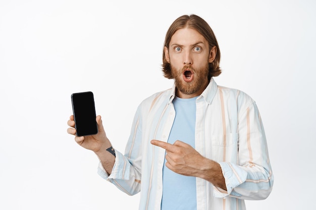 Image of bearded blond guy looks surprised, pointing finger at mobile phone screen with shocked face, big online sale, recommending download app, white background