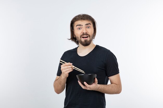 Image of bearded man holding bowl with chopsticks over a white wall.