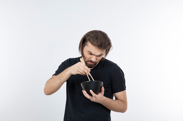 Image of bearded man holding bowl with chopsticks over a white wall.