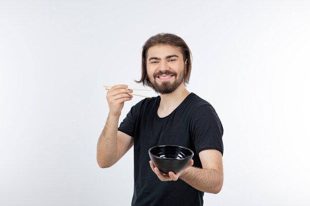 Image of bearded man holding bowl with chopsticks over a white wall.