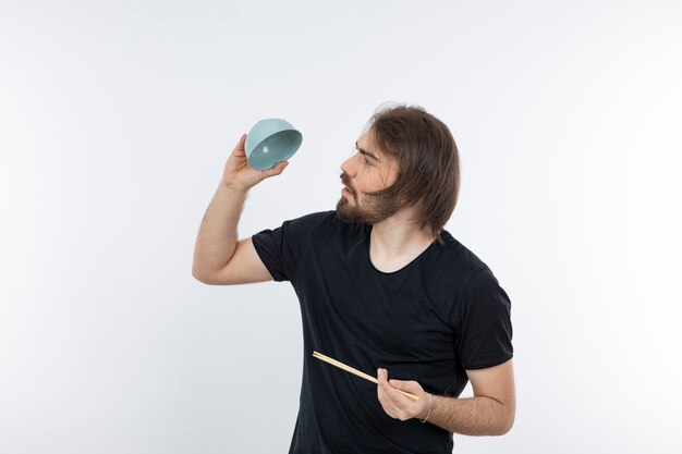 Image of bearded man holding bowl with chopsticks over a white wall.