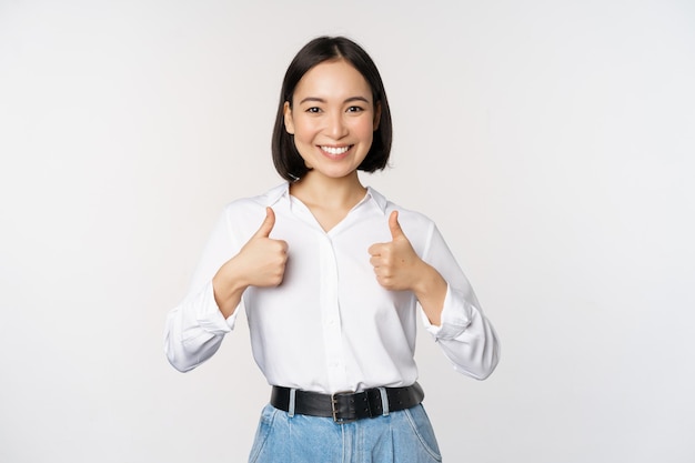 Image of beautiful adult asian woman showing thumbs up wearing formal office university clothing recommending company standing over white background