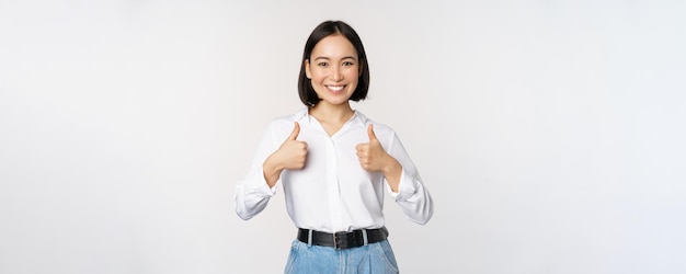 Image of beautiful adult asian woman showing thumbs up wearing formal office university clothing recommending company standing over white background