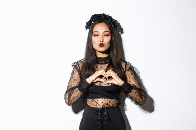 Image of beautiful asian gothic girl close eyes and pouting for kiss, showing heart gesture, express love or sympathy on halloween party, standing over white background.