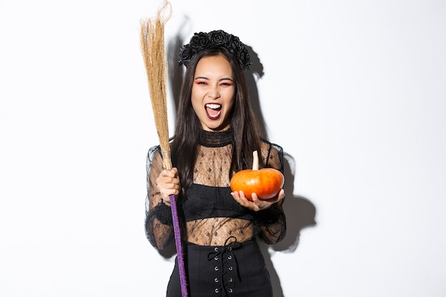 Image of beautiful asian woman dressed-up as a witch for halloween party, holding broom and pumpkin, standing over white background.