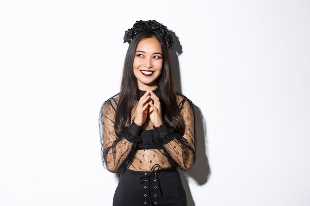 Free photo image of beautiful asian woman dressed-up as a witch or widow in black lace dress, gothic wreath, smiling pleased upper left corner and steeple fingers while prepare plan.