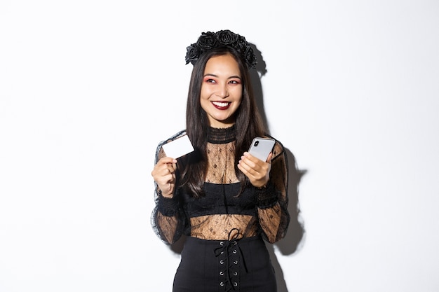 Изображение красивой азиатской женщины в готическом кружевном платье и черном венке, смотрящей в сторону приятно и усмехаясь, держа мобильный телефон с кредитной картой.