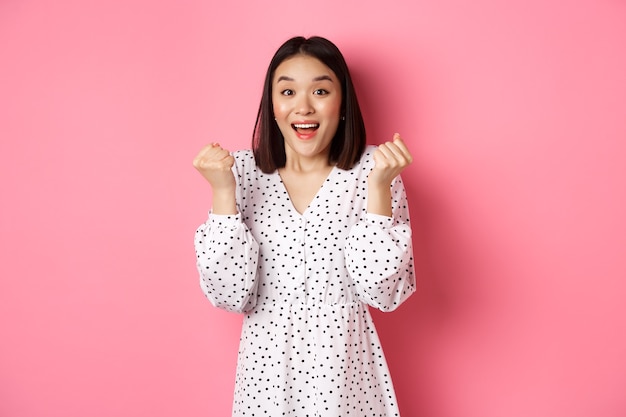 Image of beautiful asian woman winning, clench fists and saying yes with happy face, triumphing and celebrating victory, standing over pink background.