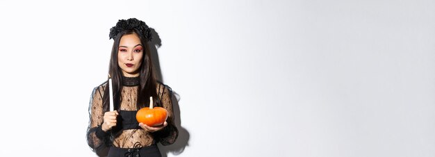 Image of beautiful asian woman in witch costume holding lit candle and pumpkin celebrating halloween