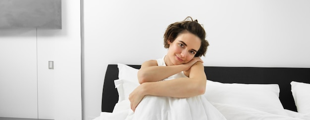 Free photo image of beautiful brunette woman sitting on bed covered in white linen sheets looking at camera