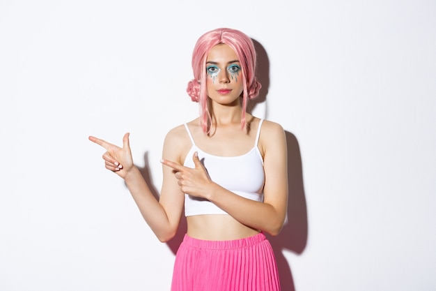 Image of beautiful caucasian girl in pink party wig and bright makeup pointing fingers left at your logo, showing banner about holidays or halloween, standing.