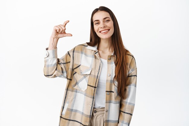 Image of beautiful caucasian woman showing holding small size thing, little gesture, smiling pleased, demonstrate product in hand, standing over white wall