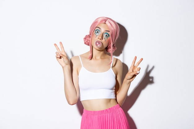 Image of beautiful girl in pink wig blowing chewing gum, popping bubble on face and showing peace signs, standing silly.