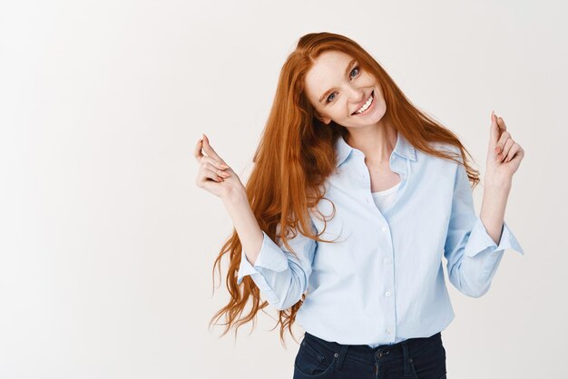 Image of beautiful girl with ginger hair and blue eyes snap fingers and dancing smiling at camera standing against white background