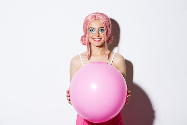 Image of beautiful glamour girl in pink wig, holding big balloon and smiling, celebrating holiday, standing.