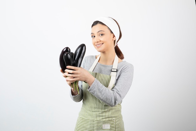 Image of beautiful happy housewife in apron holding fresh eggplants