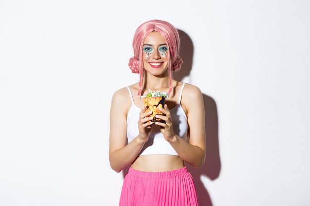 Image of beautiful smiling girl in pink wig holding trick or treat sweets, celebrating halloween, standing.