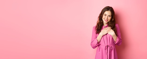 Free photo image of beautiful young woman holding hands on heart and smiling grateful express gratitude saying thank you receive heartwarming gift standing over pink background