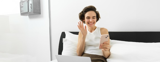 Free photo image of beautiful young woman relaxing at home sitting on bed with laptop and smartphone connects