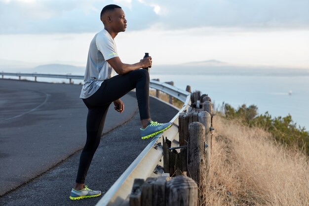 Image of black motivated man holds bottle of fresh drink, dressed in tracksuit, focused into distance, admires beautiful nature, enjoys freash air, has intensive jogging exercise outdoor alone