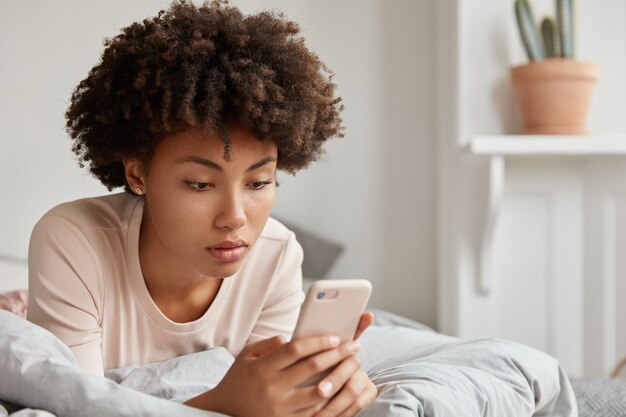 Image of black young lady with Afro haircut checks new update on social networks, shares multimedia files on mobile phone, dressed in casual nightclothes, rests in bed, makes payment online.