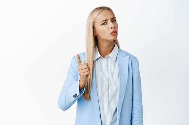 Image of blond corporate woman threatening shaking finger in disapproval scolding employee standing in suit over white background