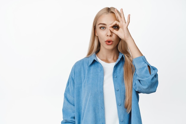 Image of blond young woman shows okay zero gesture against eye looking through fingers with impressed face expression white background