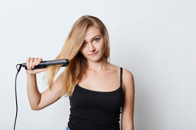 Image of blonde female in black clothing uses hair straightener, tries to do hairdo before going to party. woman does hairstyle at home, uses hair iron. female seller advertises beauty supplies