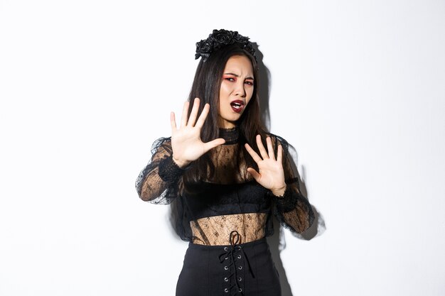 Image of bothered and annoyed asian woman in elegant gothic dress raising hands defensive, grimacing from camera flesh, asking to stop taking pictures of her, standing white background.