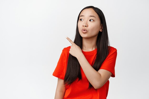 Image of brunette asian girl asking question, looking curious at upper left corner in red t-shirt on white.