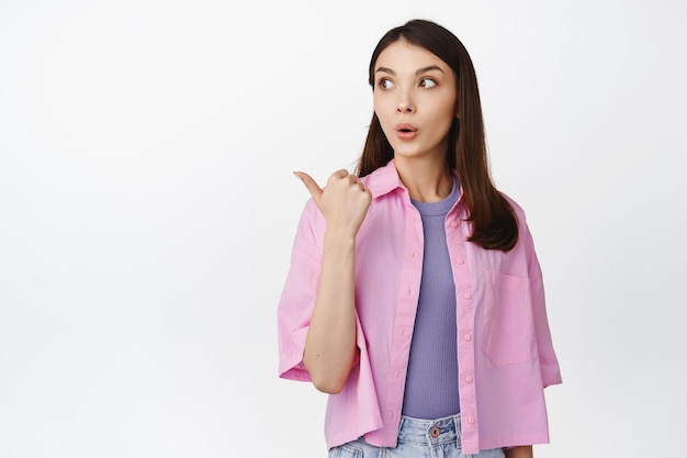 Image of brunette girl look curious at sale advertisement pointing left and looking surprised checking out promo standing over white background