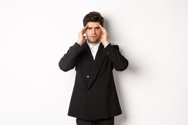 黒のスーツを着て、頭に触れて目がくらむように見え、痛みを伴う頭痛を感じ、白い背景の上に立っているビジネスマンの画像。
