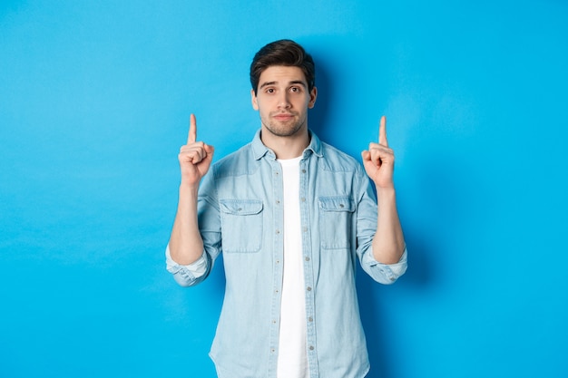 프로모션 제안을 보여주는 차분한 잘 생긴 남자의 이미지, 복사 공간에서 손가락을 가리키고 파란색 배경에 서 있는