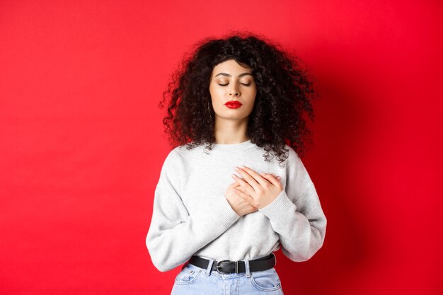 Image of calm young woman with curly hairsty close eyes and holding hands on heart keeping warm memo...