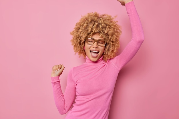 Free photo image of carefree optimistic ambitious young woman does champion dance raises hands up celebrates something wears spectacles and turtleneck isolated over pink background triumps over success.