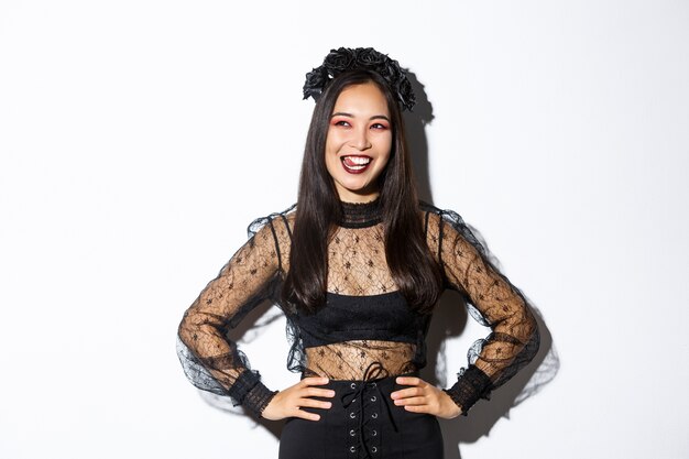 Image of carefree smiling asian woman in gothic dress and black wreath looking thoughtful at upper left corner, licking lips from desire or temptation, standing over white background.