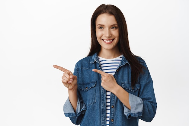 Immagine di una donna caucasica che punta, guardando a sinistra con un sorriso soddisfatto, una cliente femminile punta sul prodotto che desidera, raccogliendo qualcosa in negozio, in piedi su sfondo bianco.