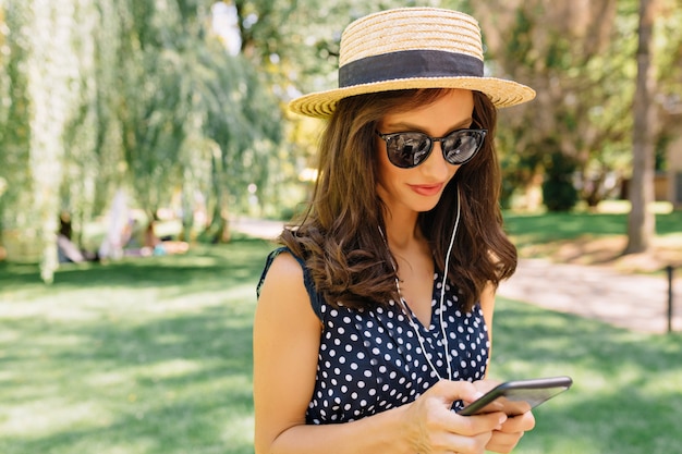 Image of charming style woman is walking in the summer park wearing summer hat and black sunglasses and cute dress.