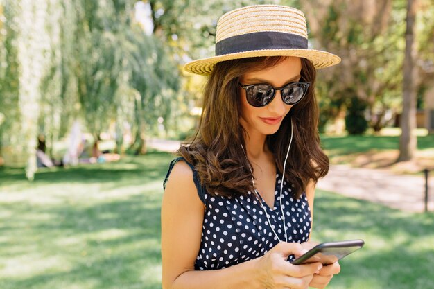 Image of charming style woman is walking in the summer park wearing summer hat and black sunglasses and cute dress.